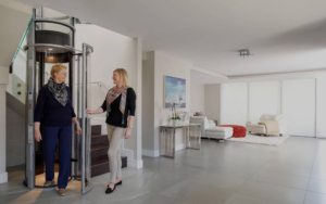 Two women talking near a modern residential elevator in a bright living room.